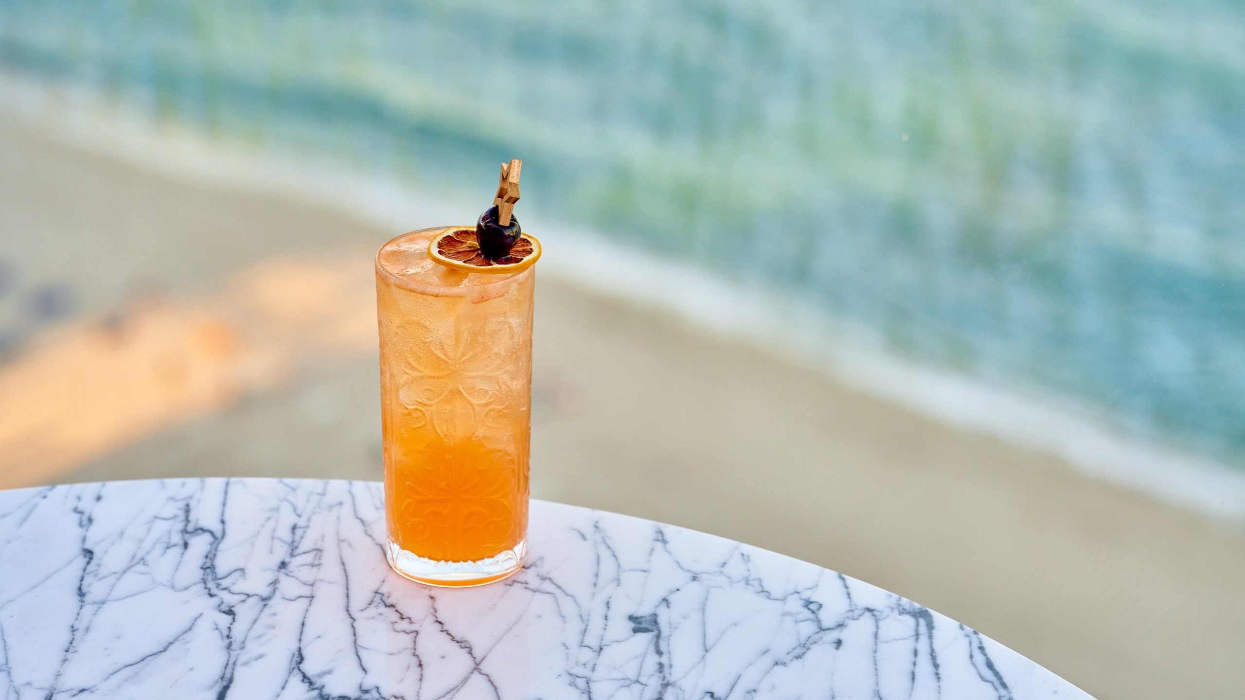 an orange cocktail with a a garnish of a dried orange slice upon a marble table