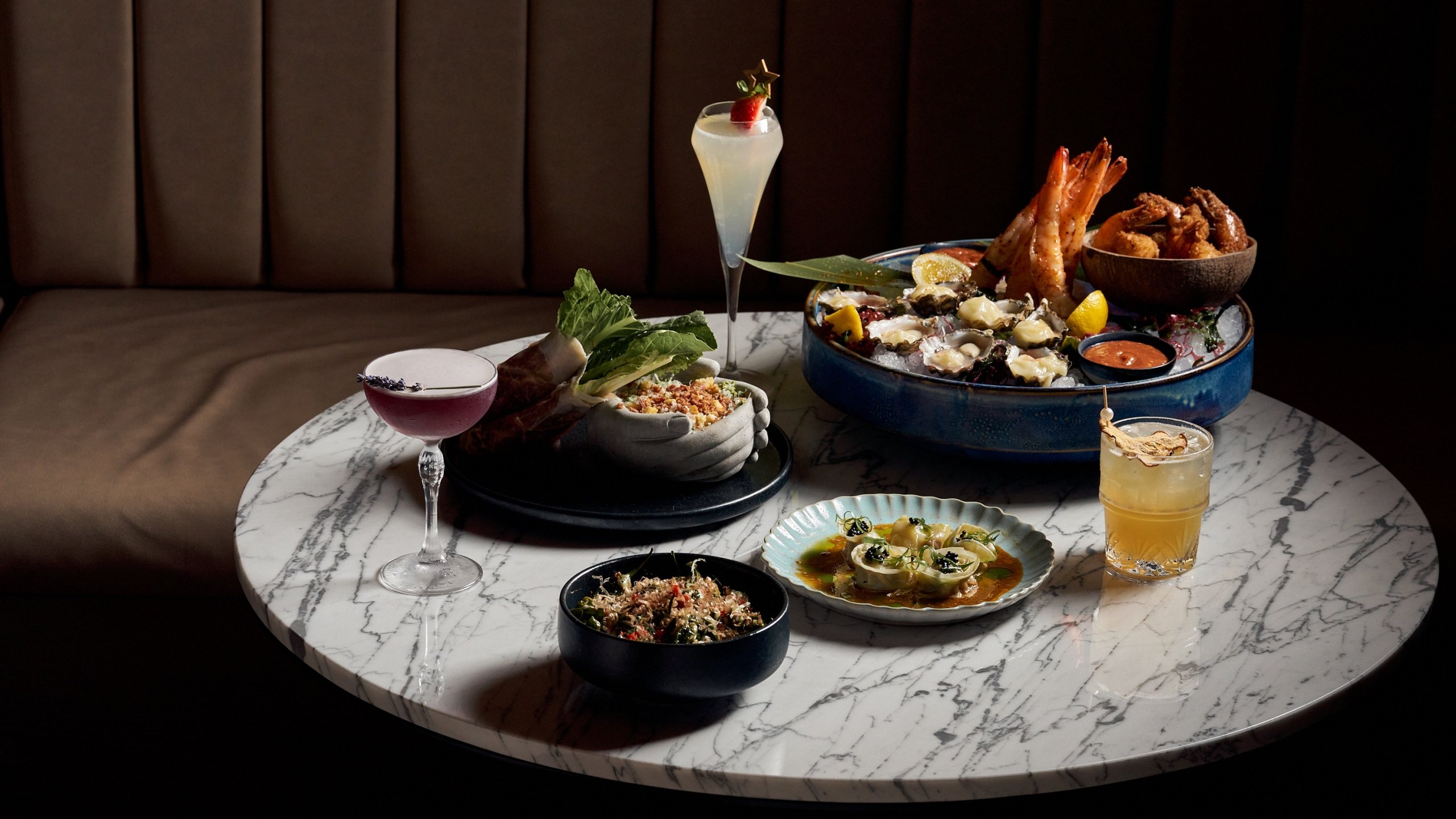 three cocktails and three small plates of food next to a seafood plater on a marble table