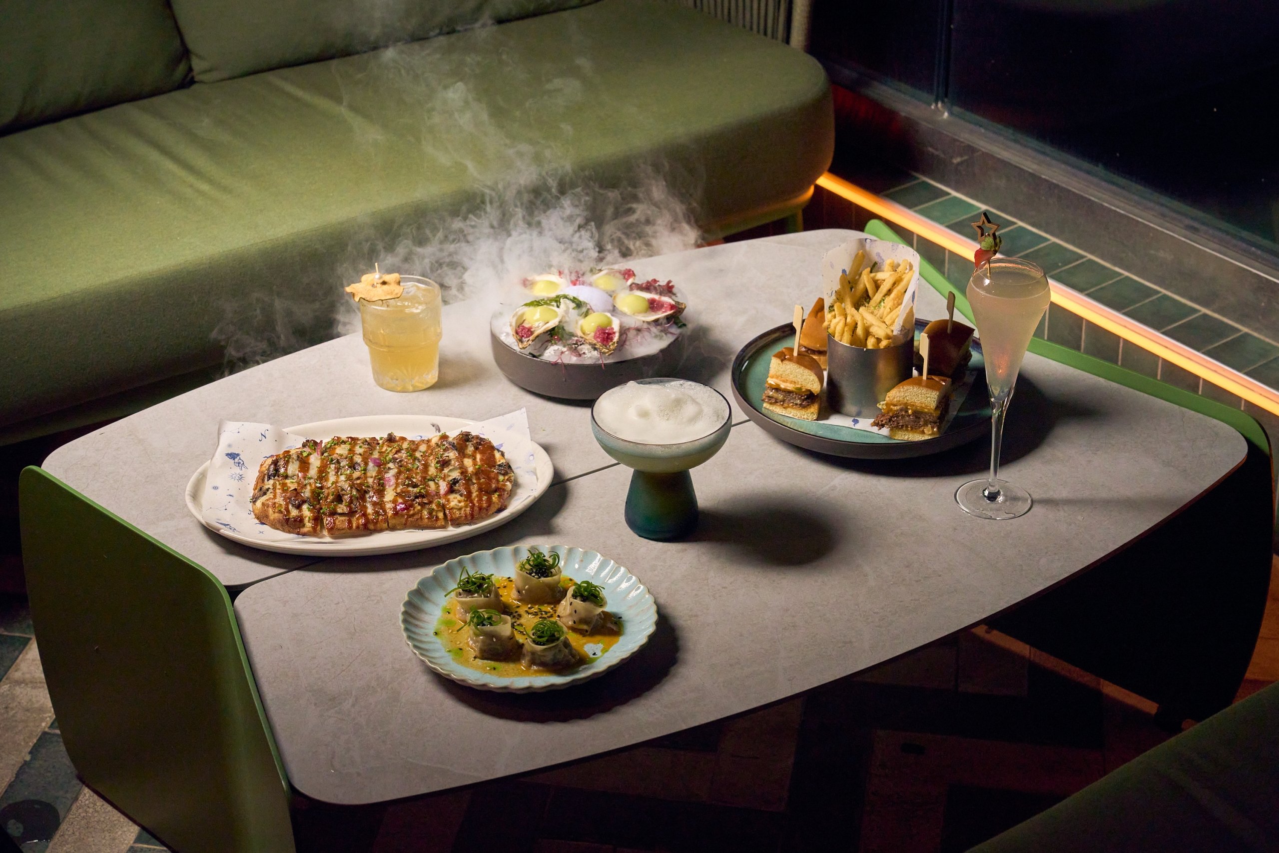 various dishes and cocktails on a table with a green bench in the background as steam rises from a dish