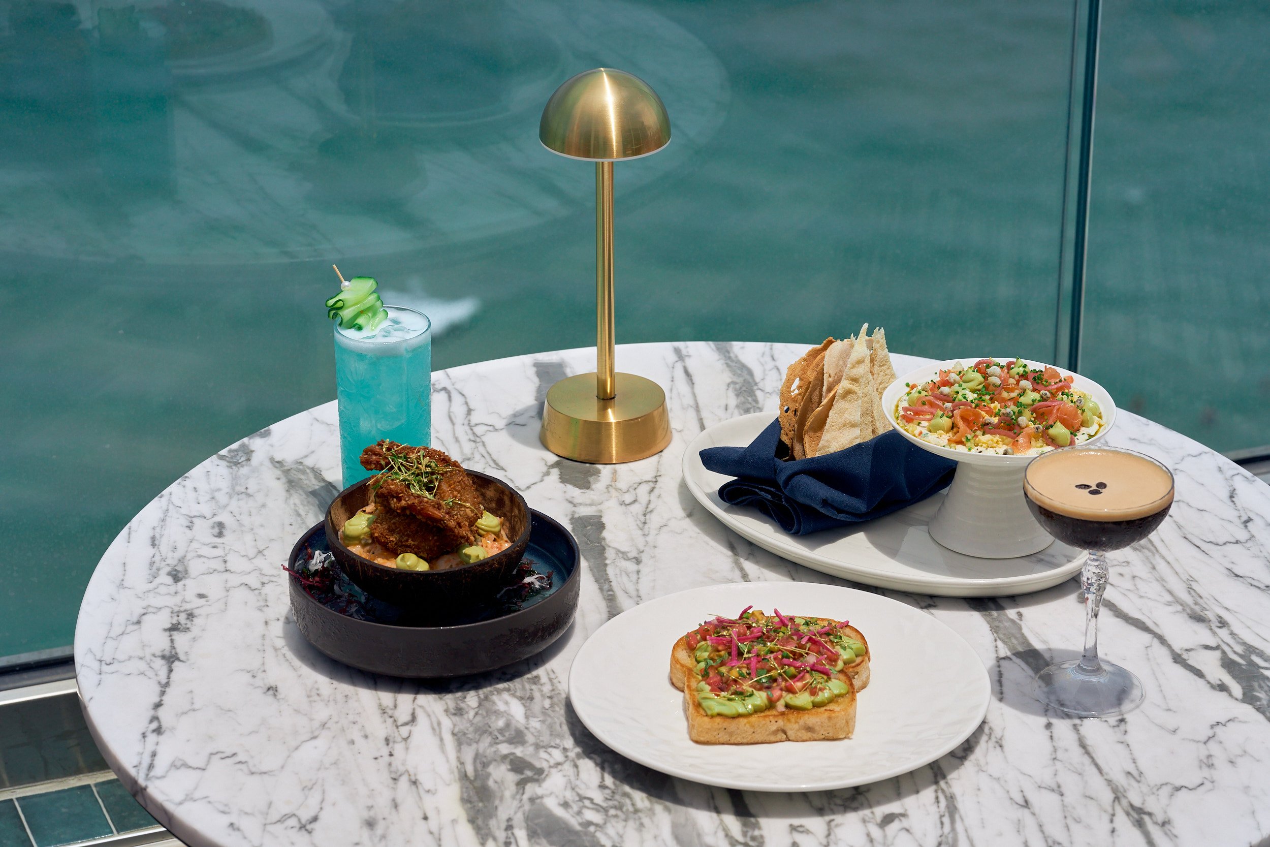 a blue cocktail and coffee cocktail next to a dish of avocado toast, a colorful dip and a fried chicken dish