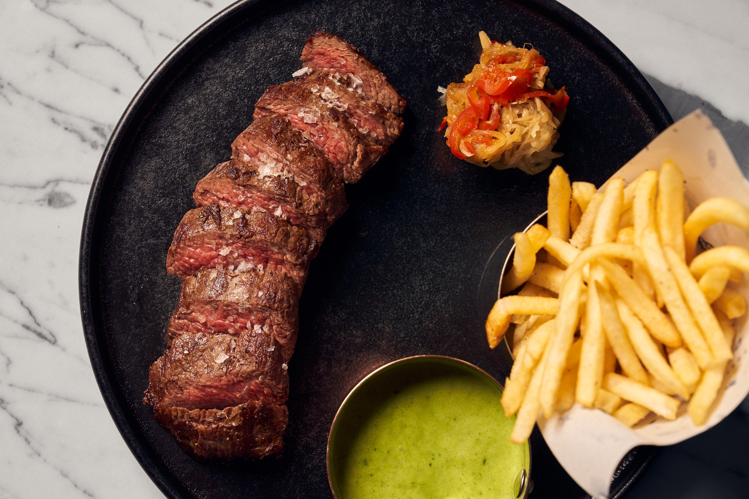 sliced steak, a green dipping sauce and a basket of fries upon a black plate
