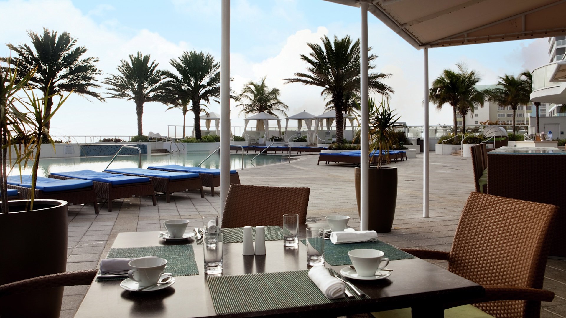 View from patio at Ilios Greek restaurant. Brown chair next to dark wood tables and green place settings in view of the pool