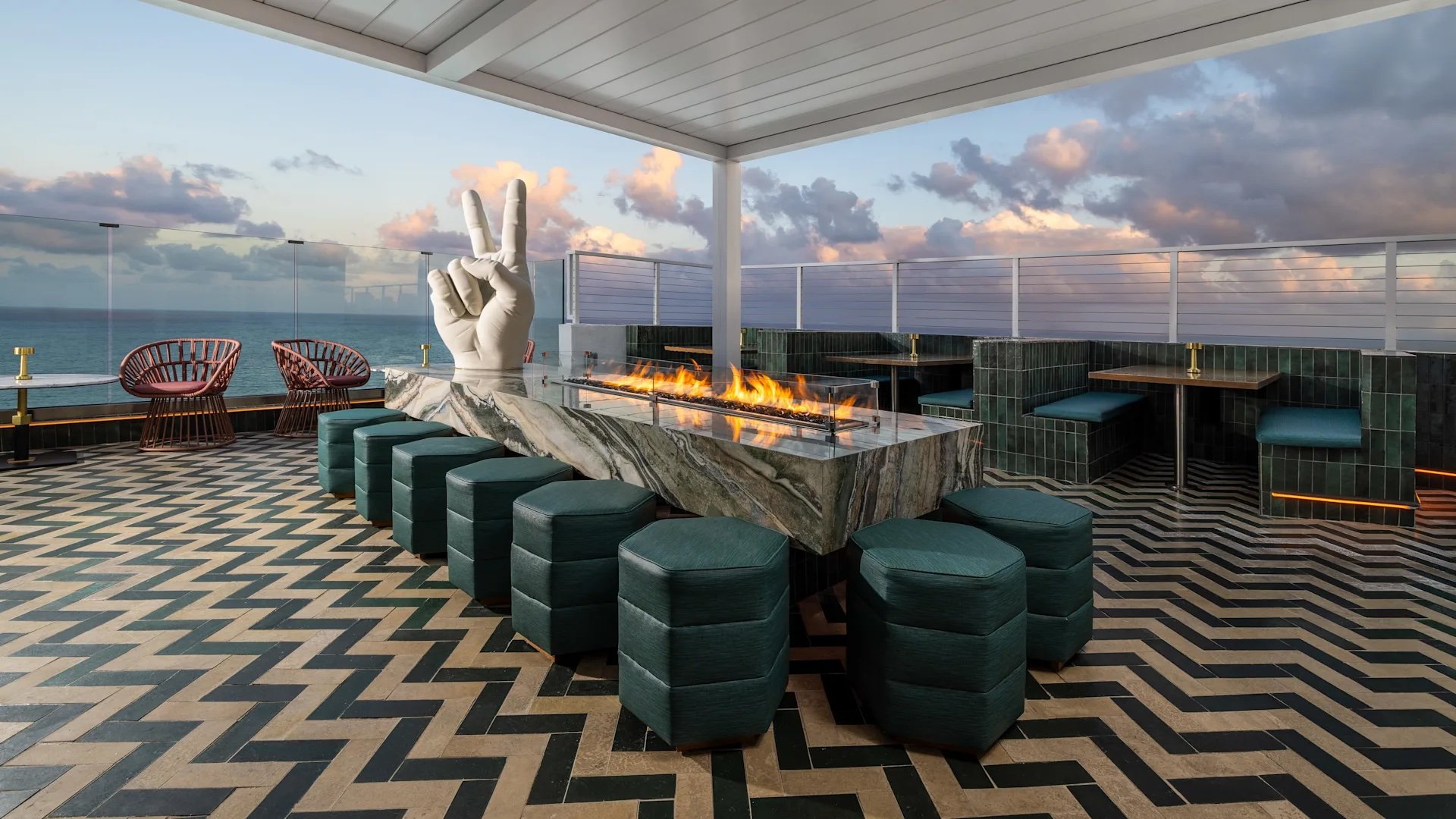A rectangular marble table contains a a gas fire pit protected by glass, surrounded by padded dark green stools