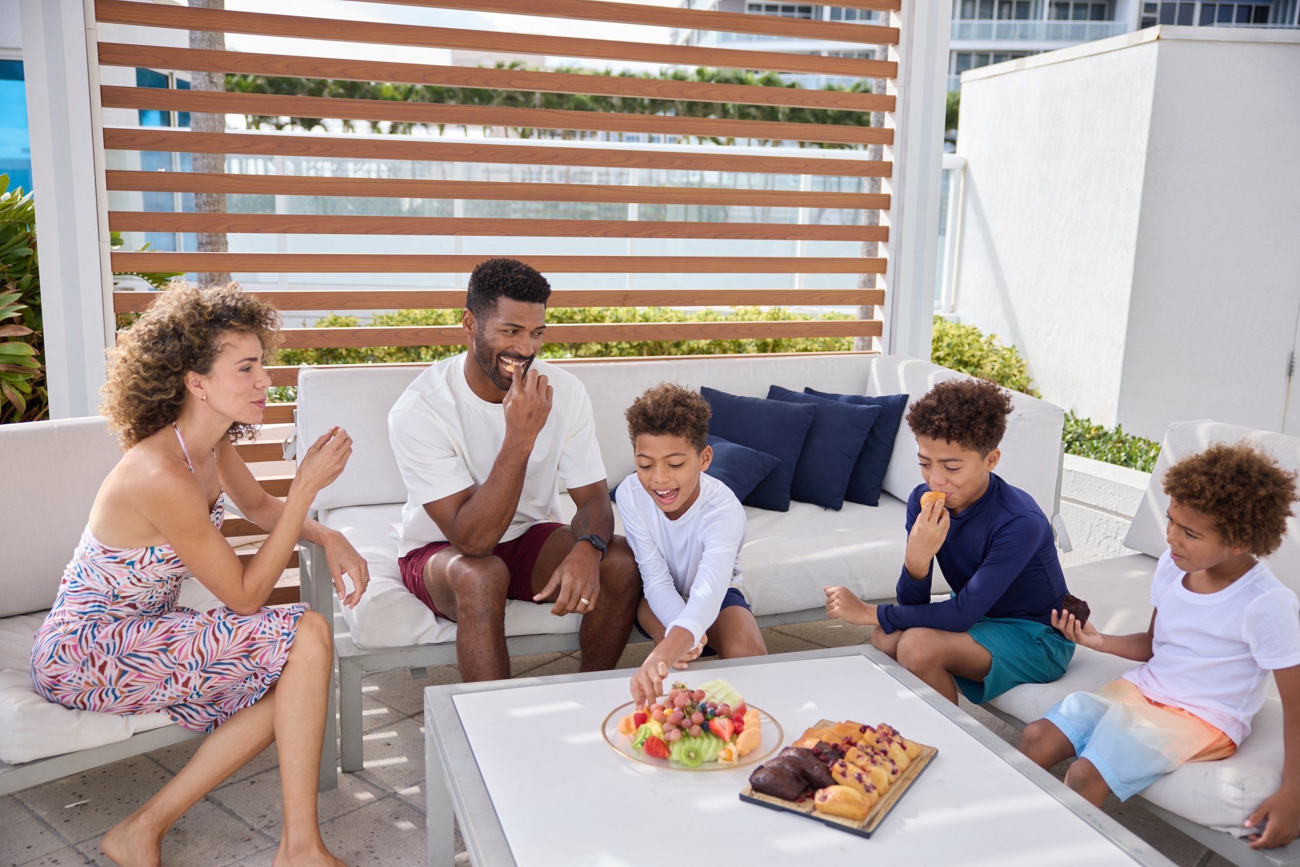 Family of five enjoying fruit and other snacks on a patio, sitting on white exterior furniture