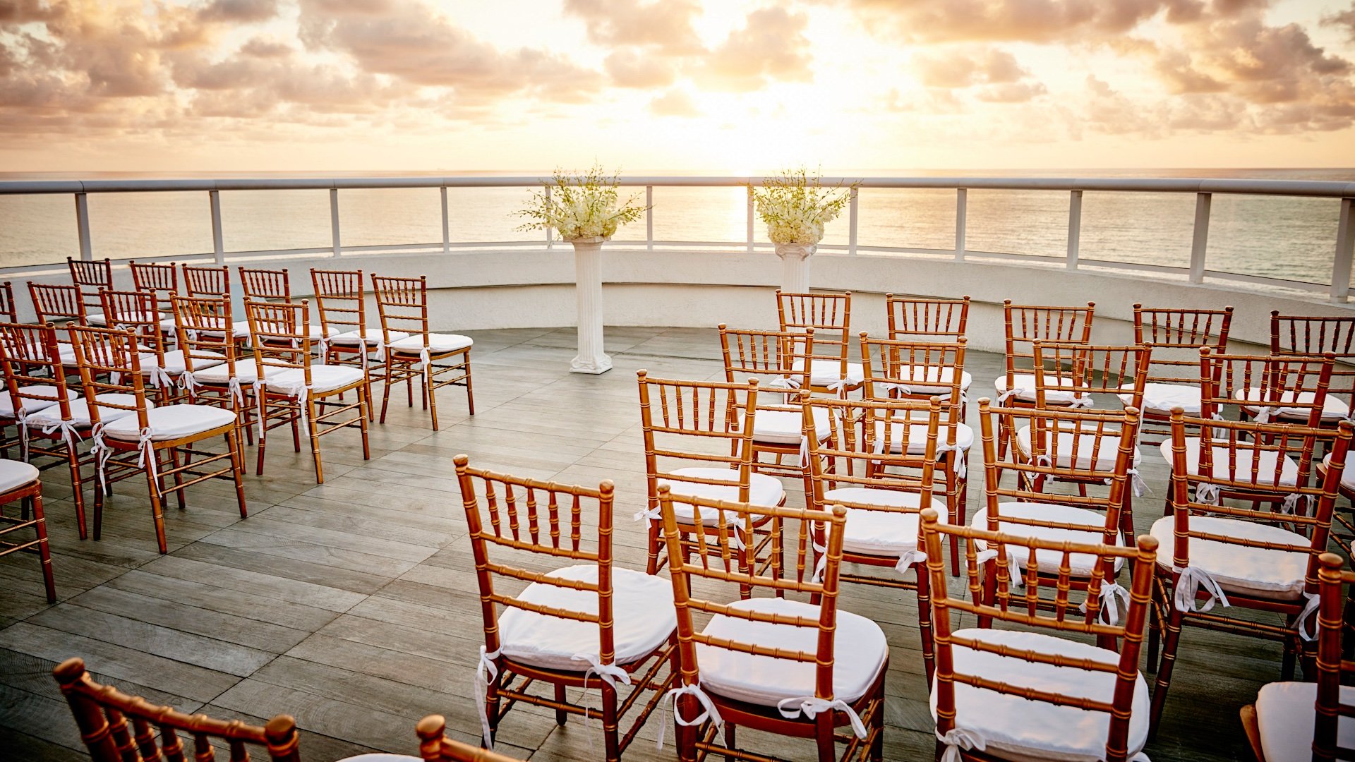 wooden chairs with white cushions line a path towards two freestanding columns adorned with flowers at sunset