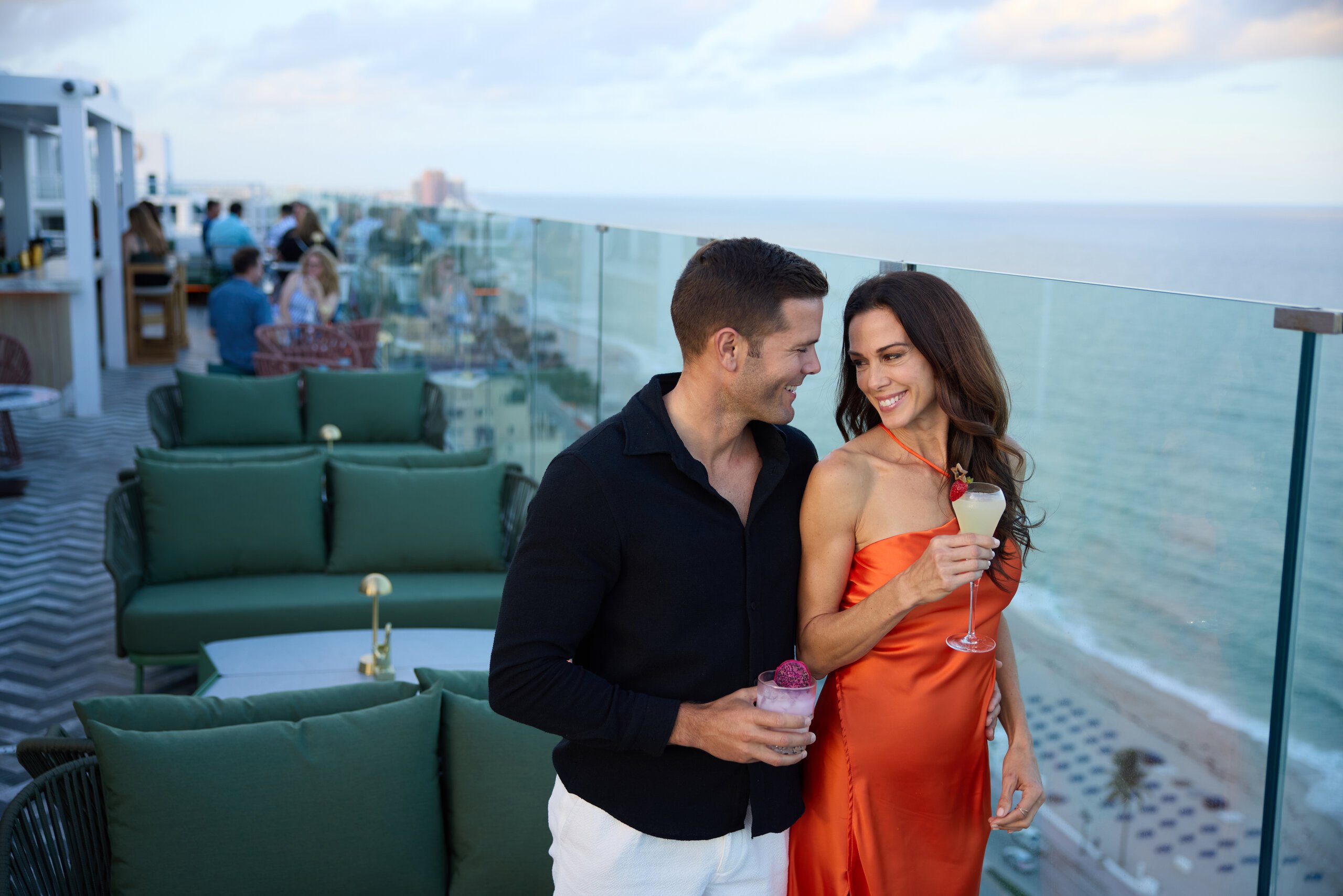 Couple holds cocktails at Nube Rooftop restaurant while they smile at each other.