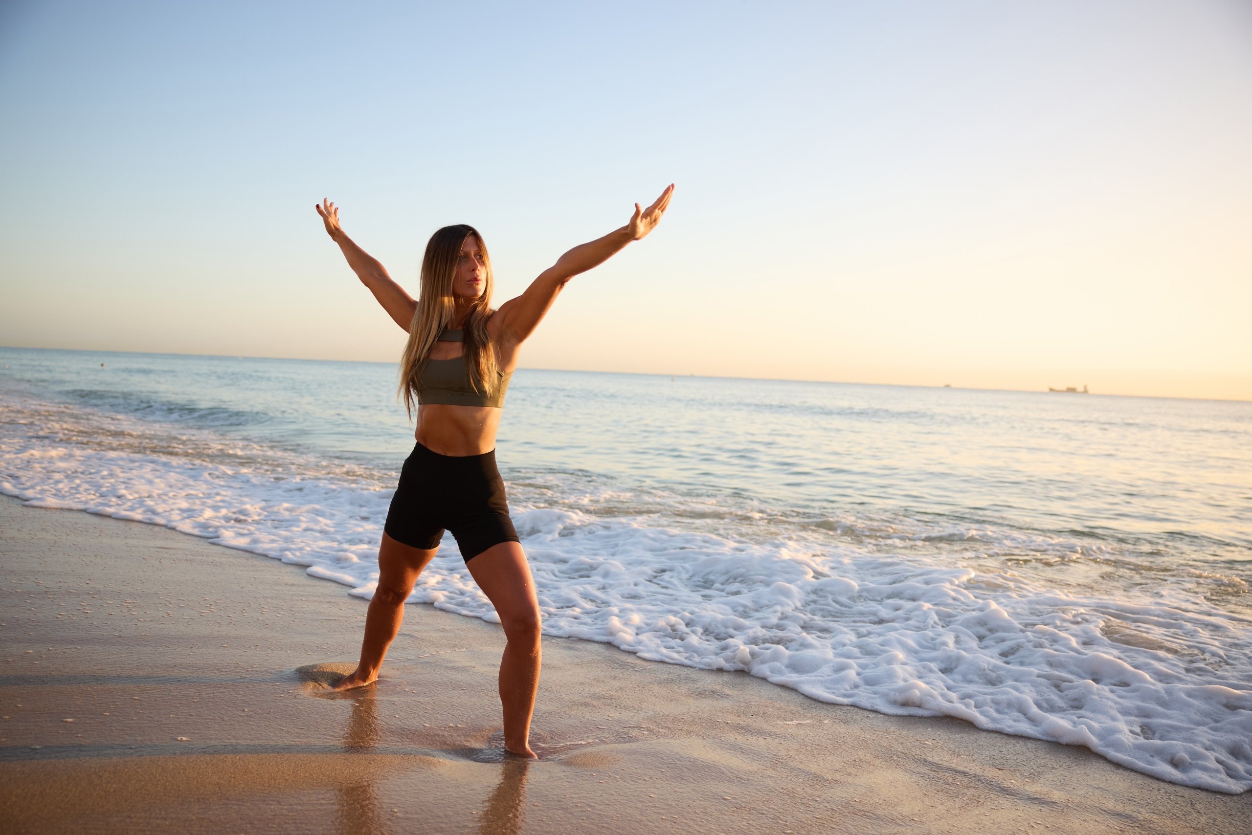 Woman in a green top and black short performs yoga on the beach as a wave recedes from her feet.