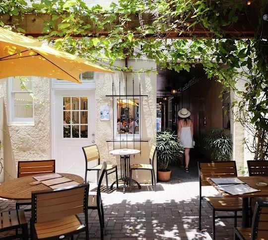 A woman in a white dress and sun hat walks down a beautiful alley, away from an outdoor dining area