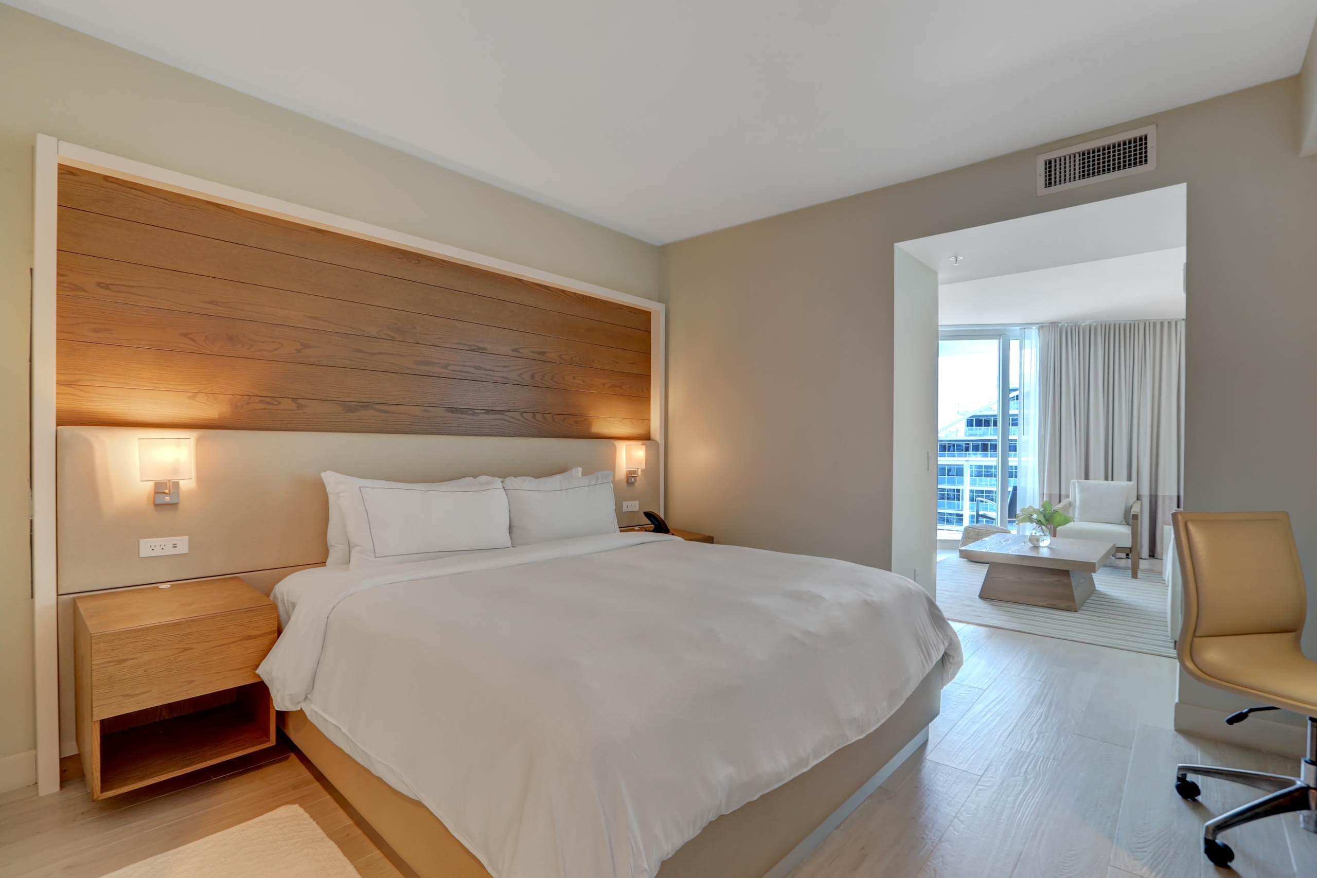 A king bed with white sheets in bedroom connected to living room with large window to balcony