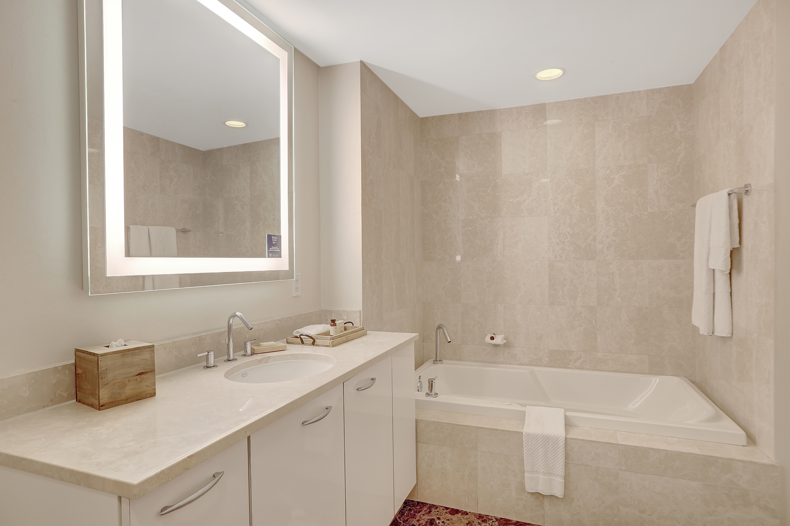 Sink counter with hotel amenities and a bright mirror and white bathtub in marble bathroom