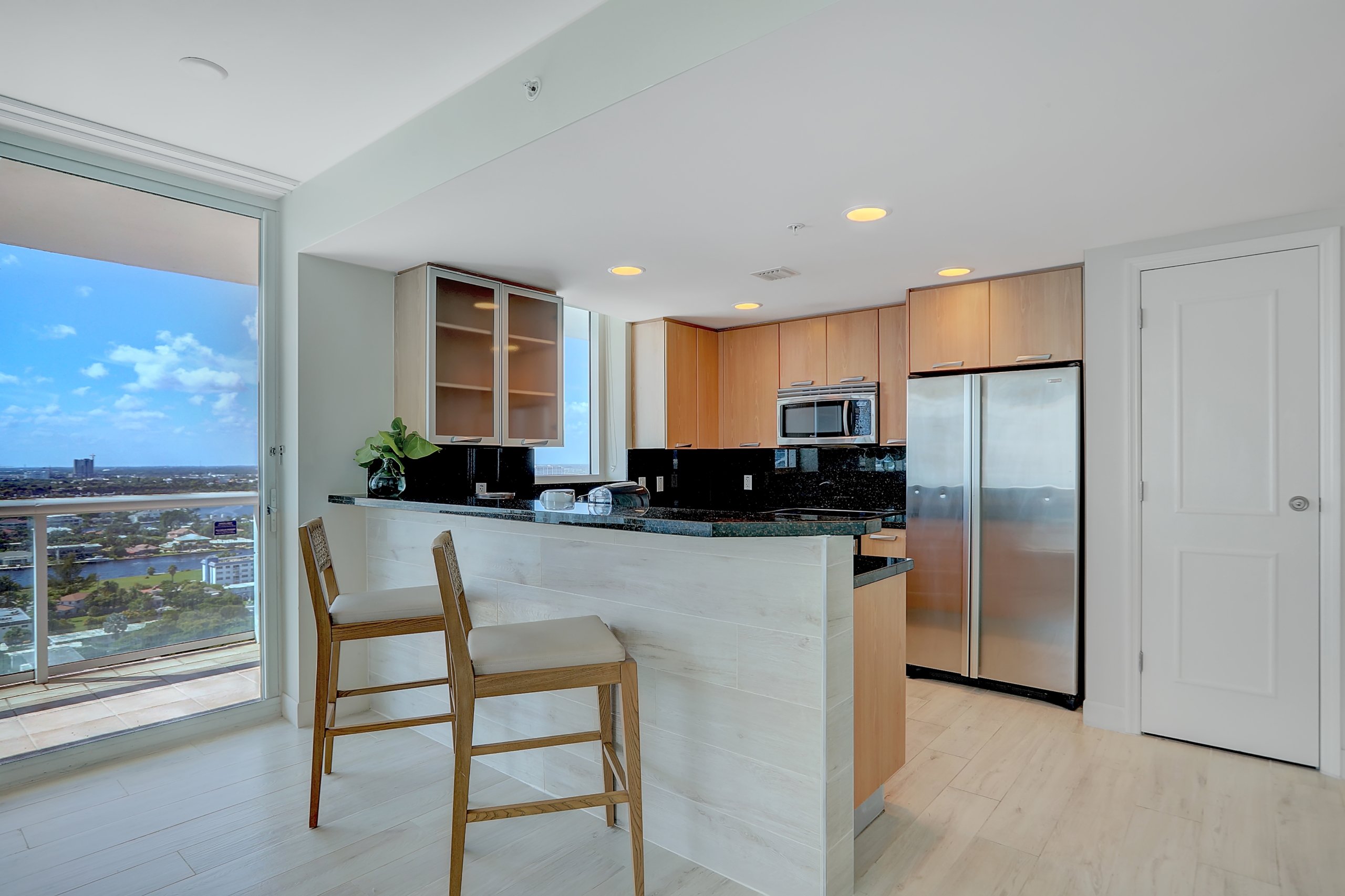 Wooden kitchen area with black counter, wooden shelves, island table and wooden tall chairs