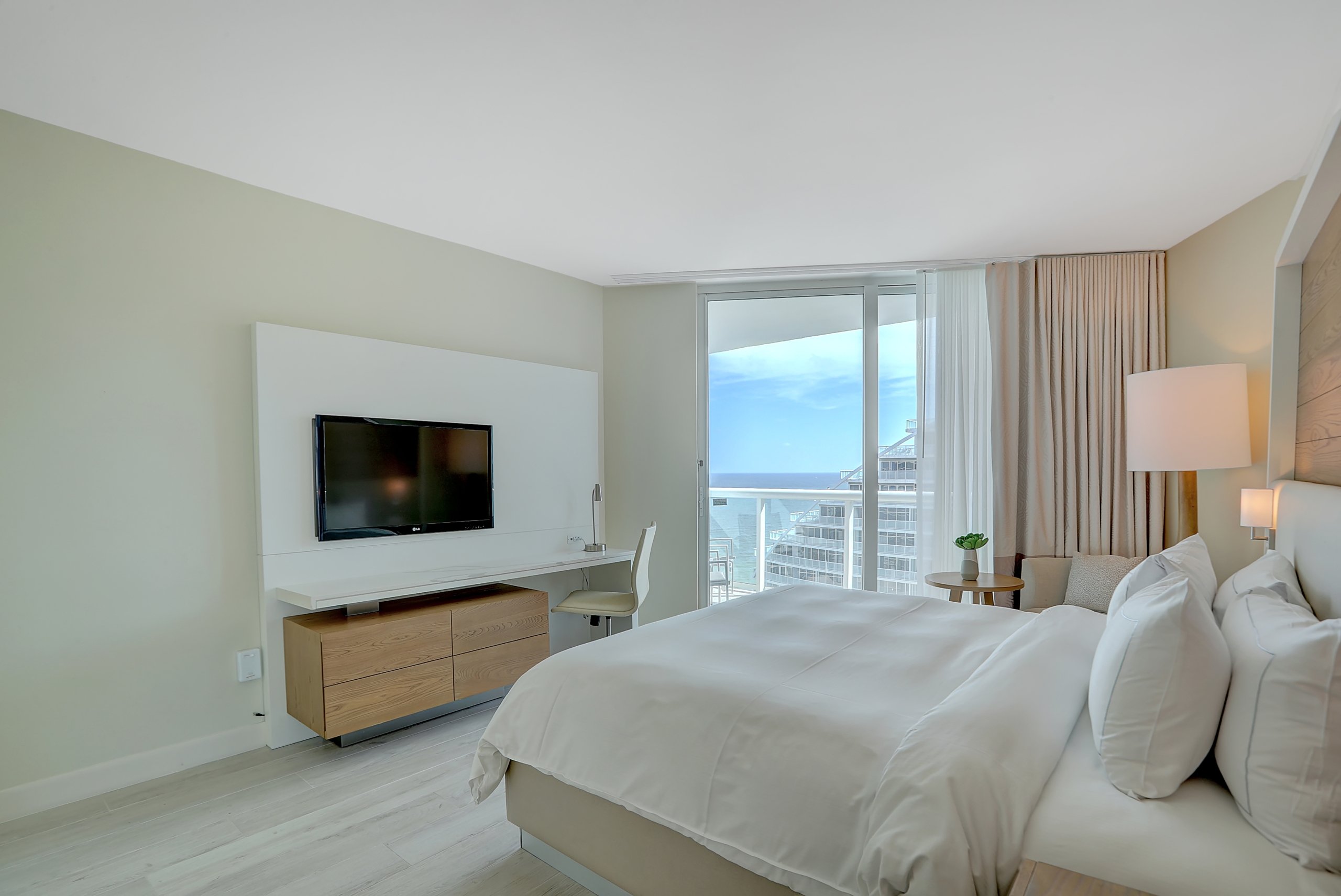 A bed with white sheets, modern chair and desk with large window to balcony in bedroom