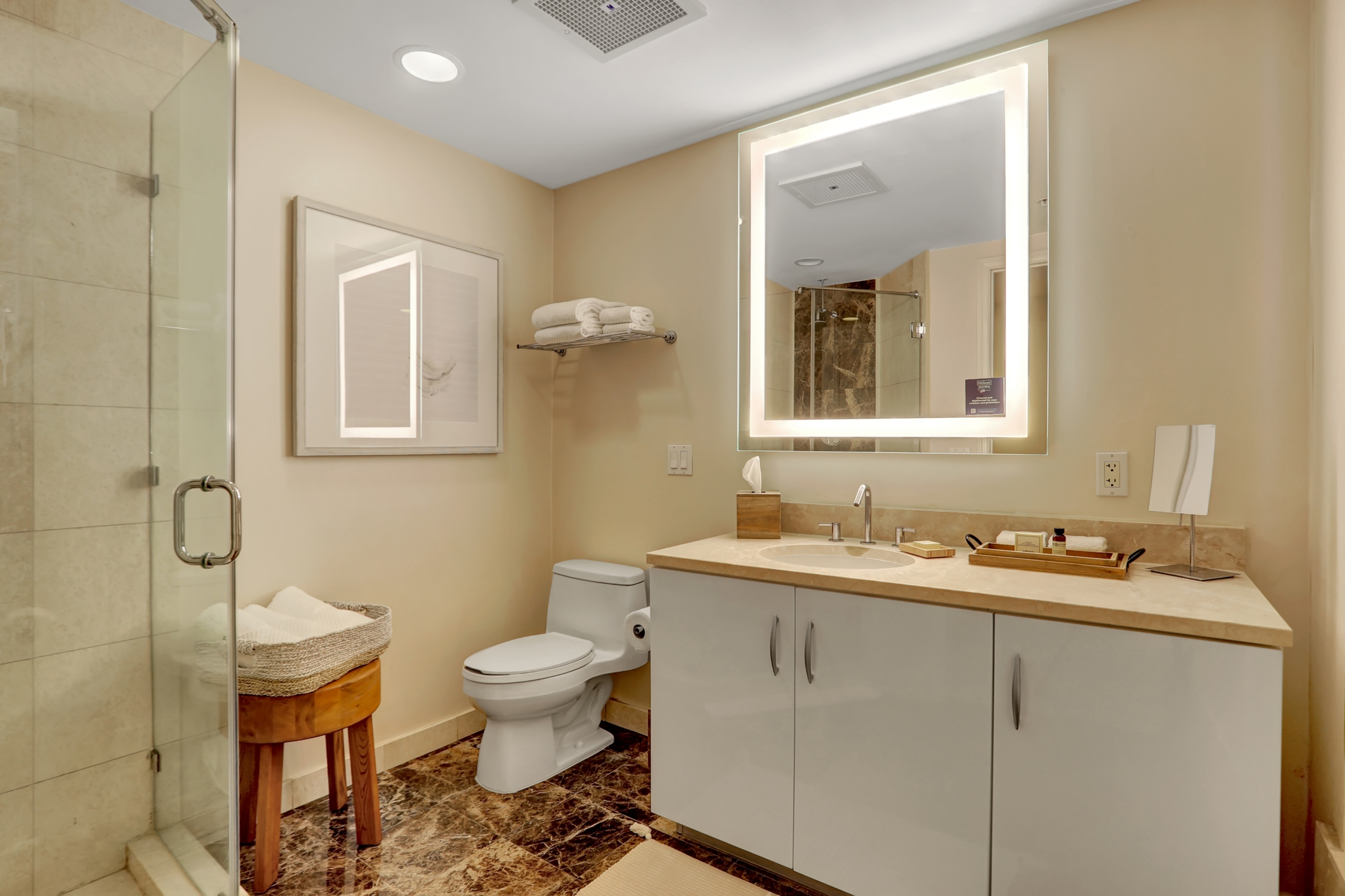 Sand colored marble bathroom with shower booth, sink counter with hotel amenities, large bright mirror