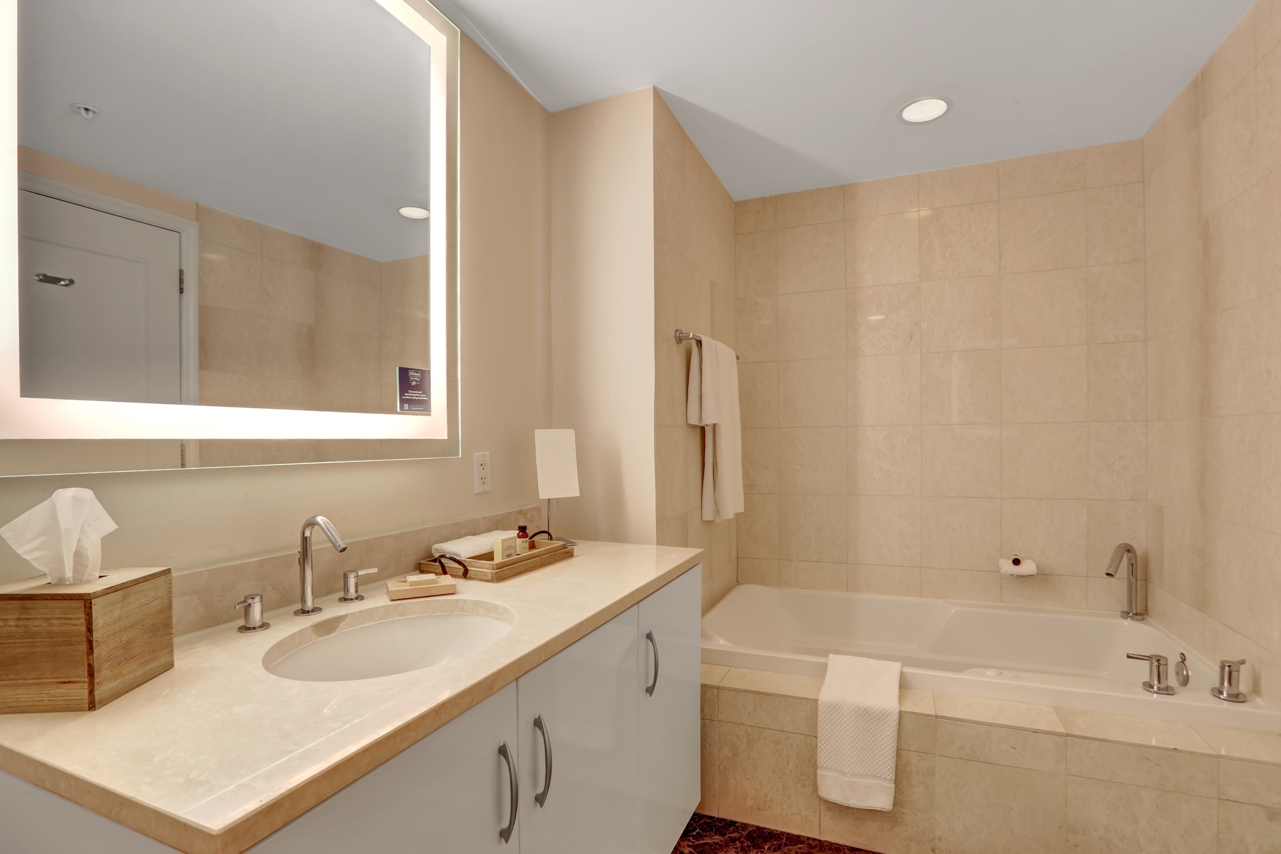 Sand colored marble bathroom with white bathtub, sink counter with hotel amenities, and large bright mirror