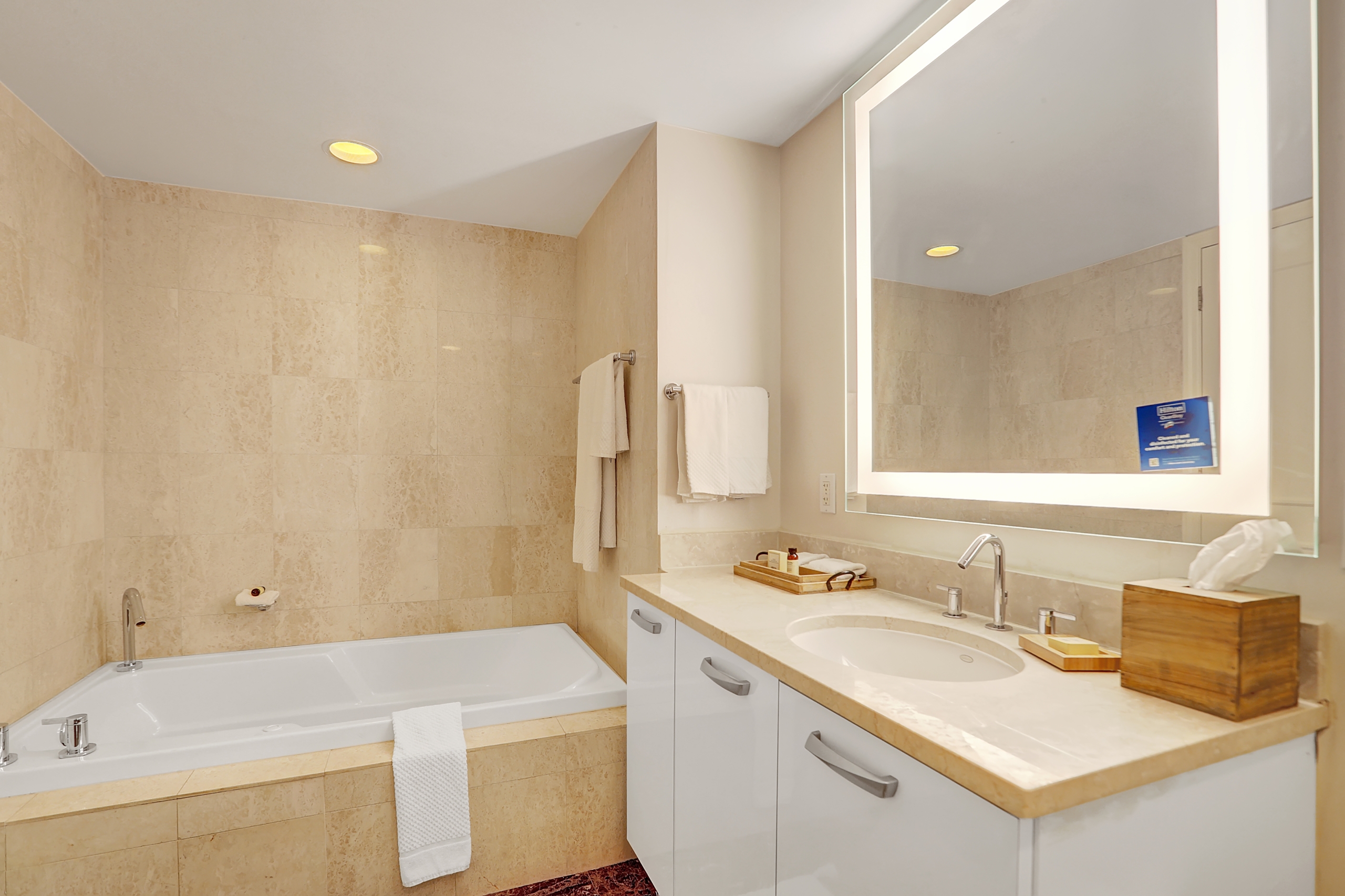 Beige marble bathroom with white big bathtub and sink counter with hotel amenities