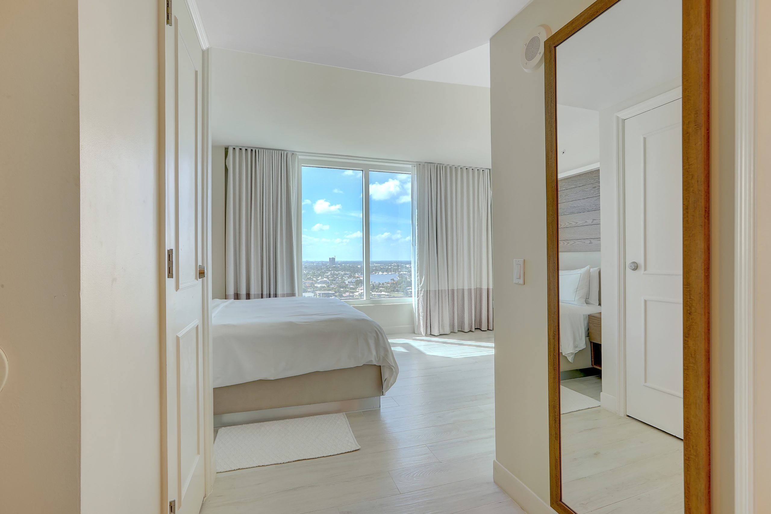 Modern white room with king bed in white sheet and huge window of city view next to tall mirror