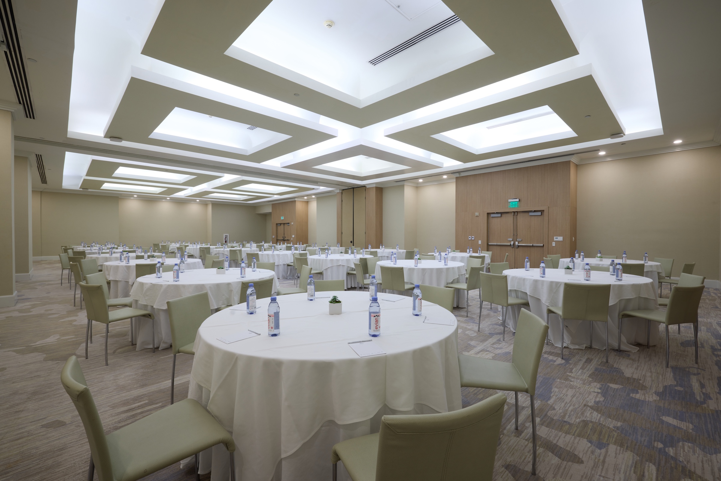 A large ballroom filled with banquet tables under a ceiling-wide geometric white light fixutre