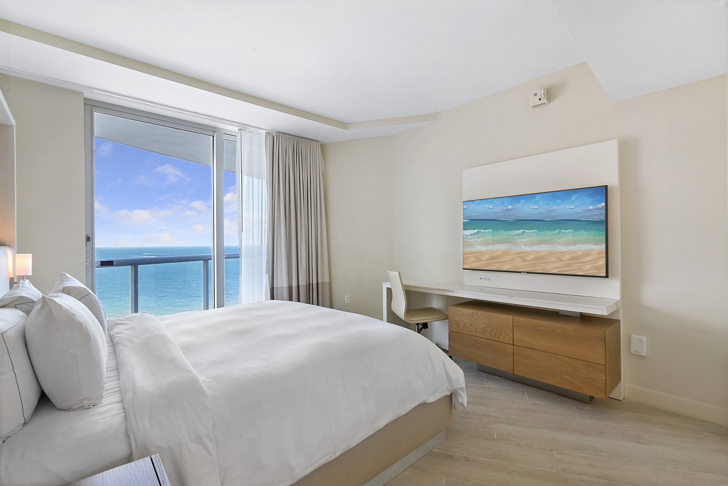 A bright bedroom with an adjacent balcony overlooking the ocean and a wall mounted television