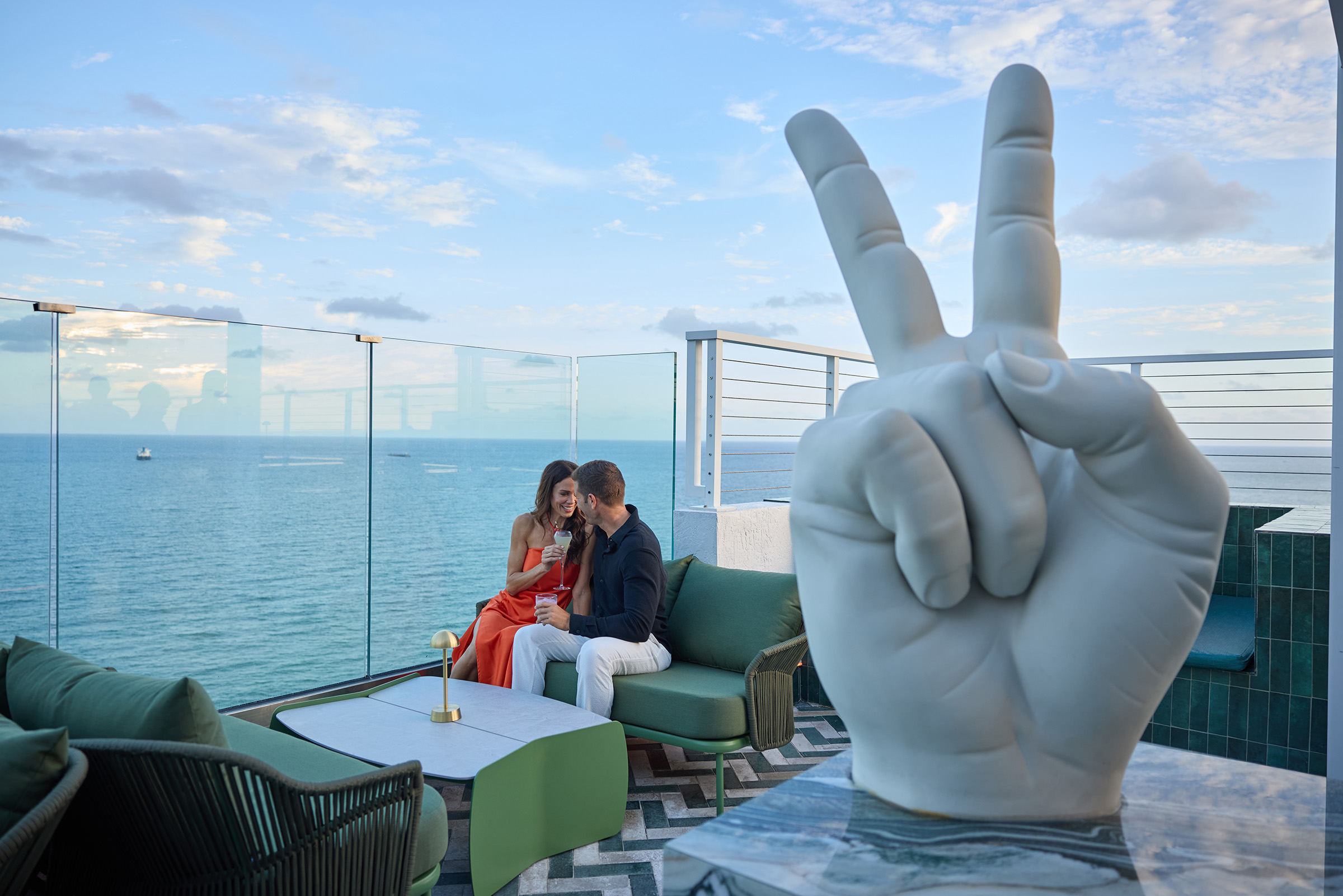 A couple enjoying their cocktails and their company with ocean view behind the V shaped statue at Nube