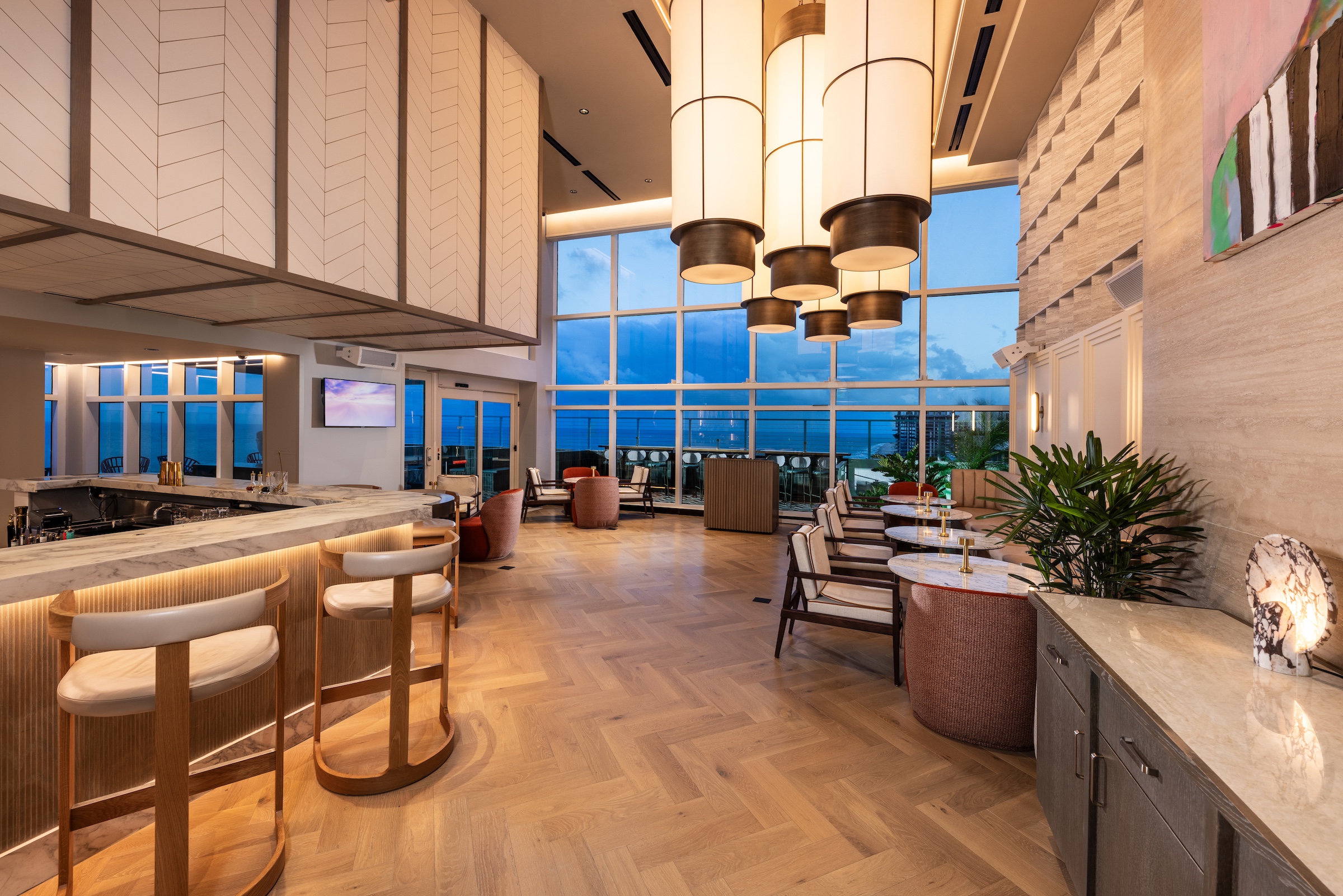 A bar and restaurant dining room decorated with large white pendant lights, with a large bay window looking out on the ocean