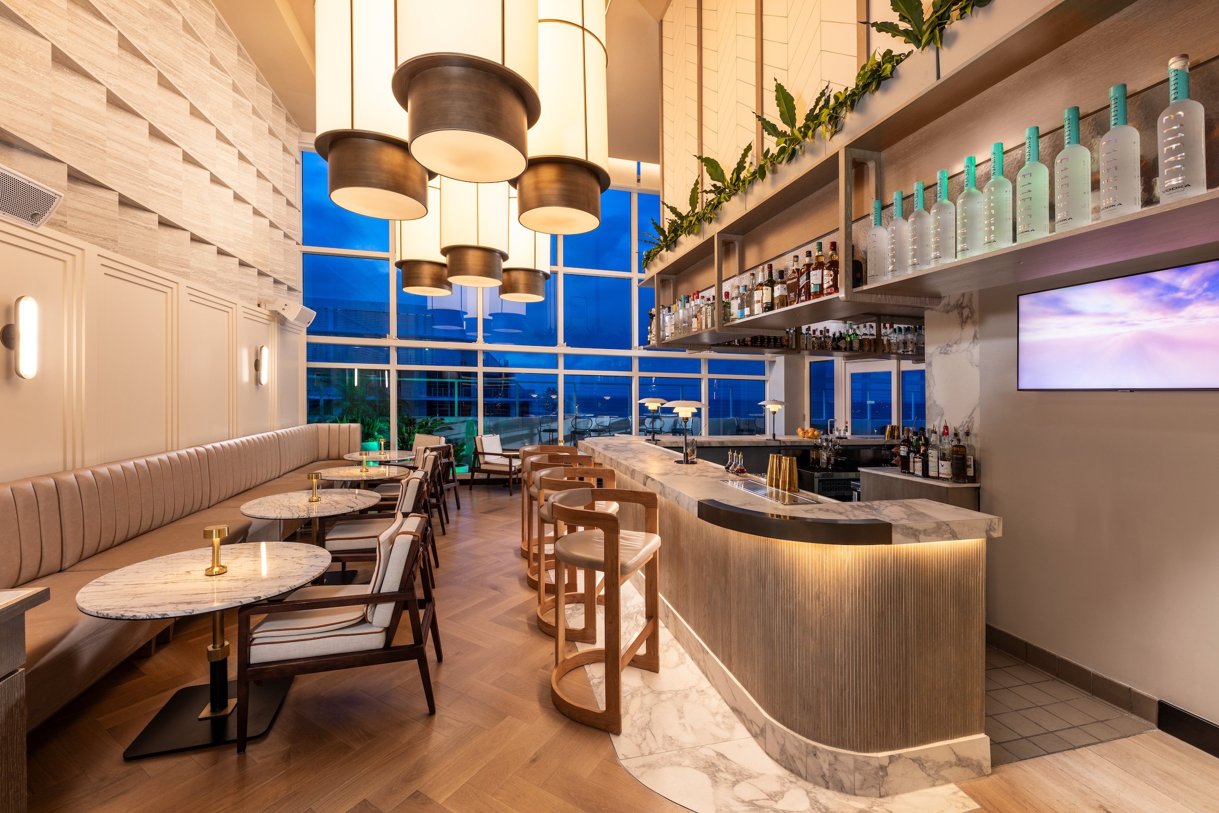 A long restaurant dining room with a bench and tables to one side, and a corner bar to the other, adjacent to a large bay window looking out on the ocean