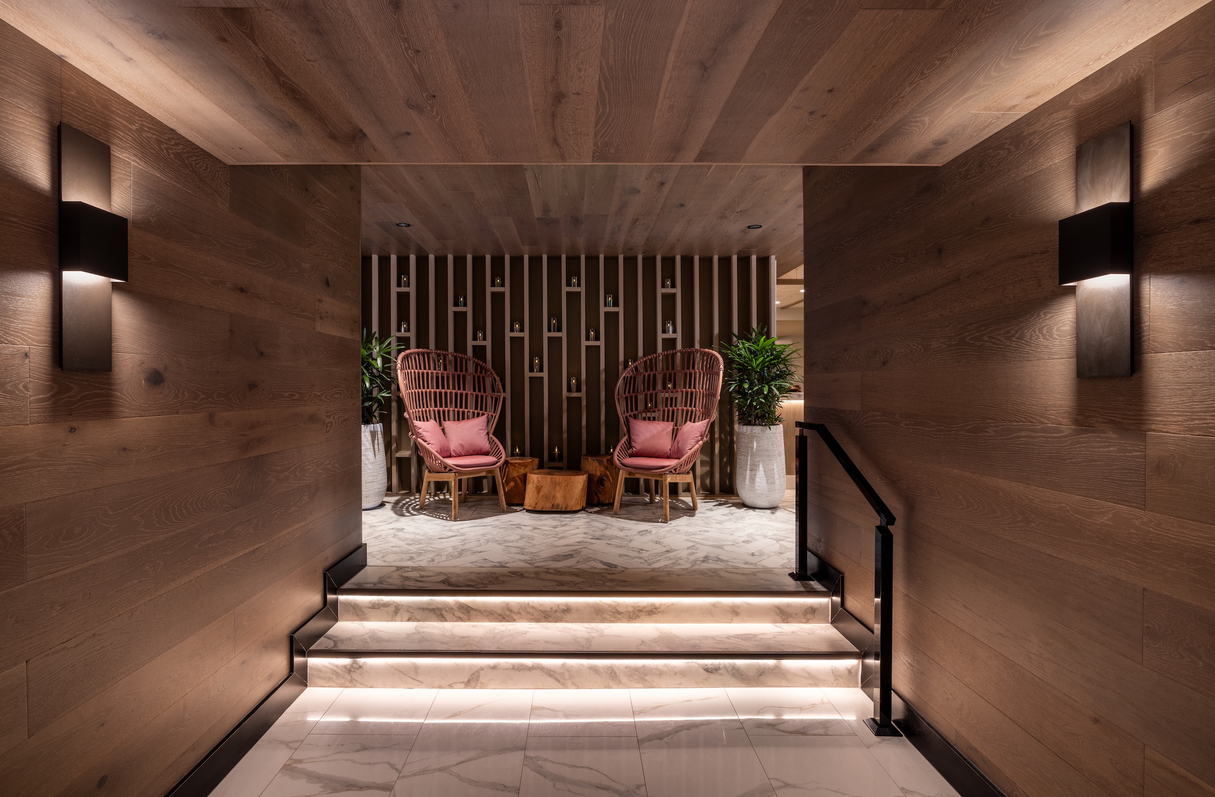 A corridor with stairs leading to two wicker armchairs flanked by tall potted plants