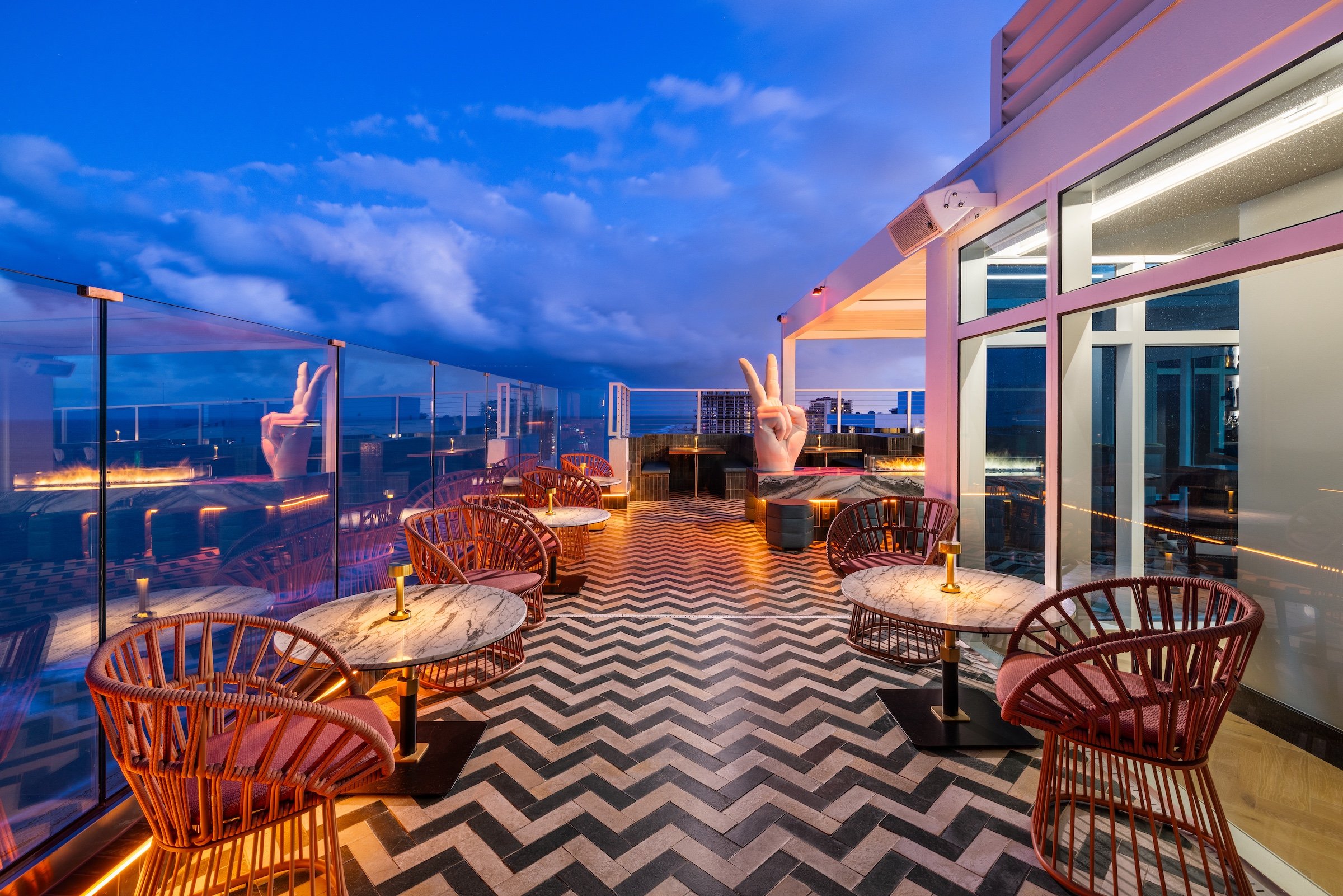 A patio looking out on the ocean at dusk, with a zigzag floor pattern and marble tables surrounded by wicker chairs