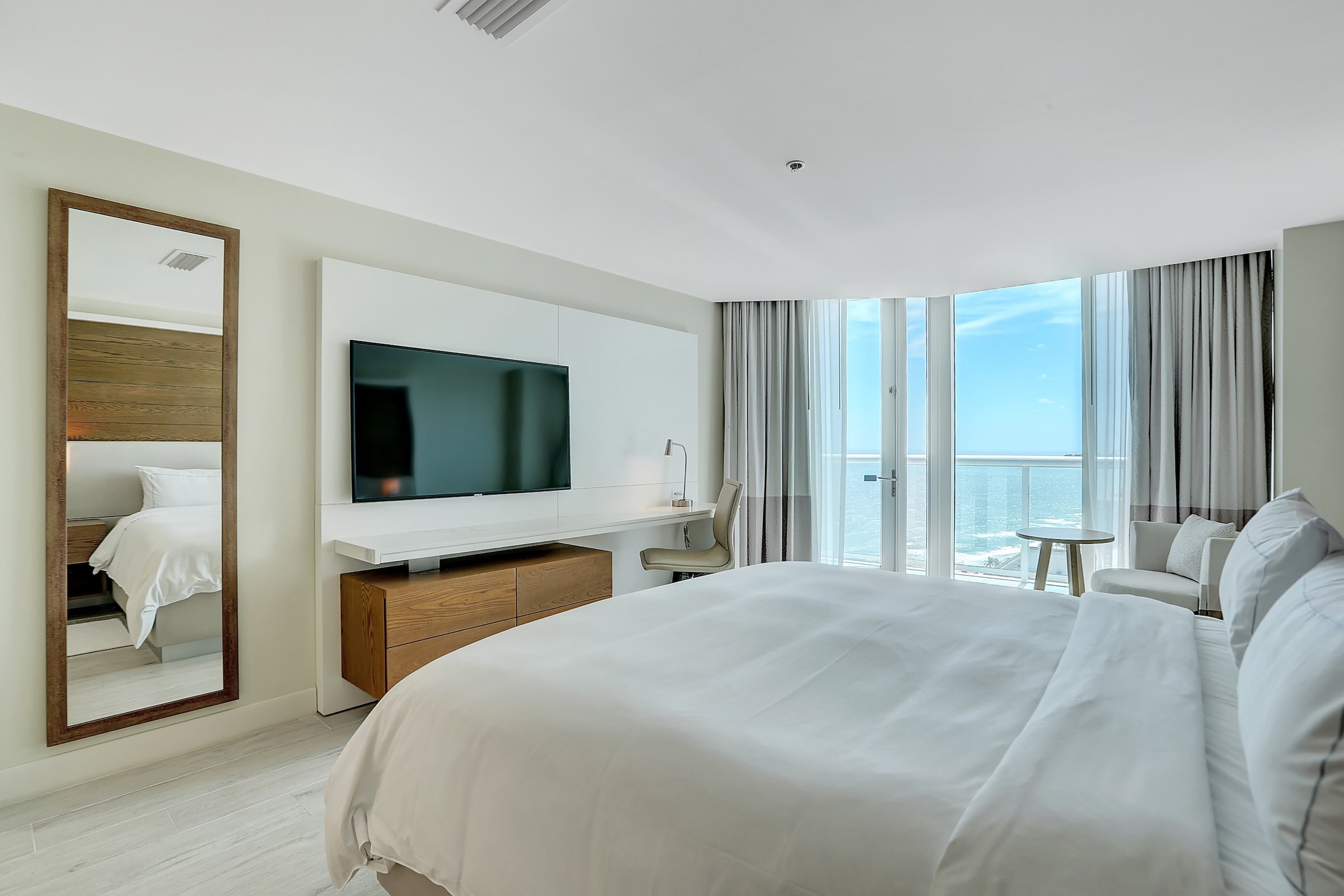 A bedroom with a large King bed opposite a wall mounted television and desk, with an adjacent balcony overlooking the ocean