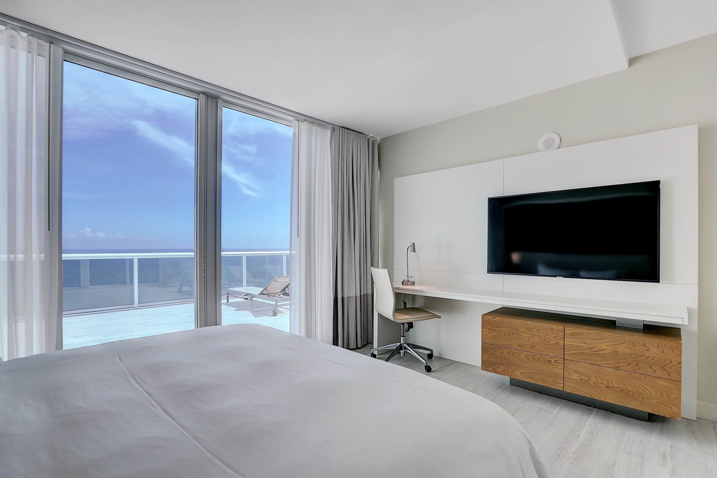 A bedroom with a large King bed opposite a wall mounted television and desk, with an adjacent balcony overlooking the ocean