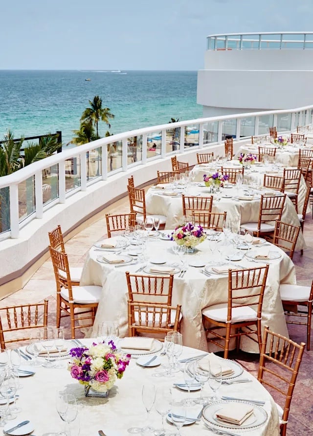 A seaside balcony with several banquet tables decorated with white linens