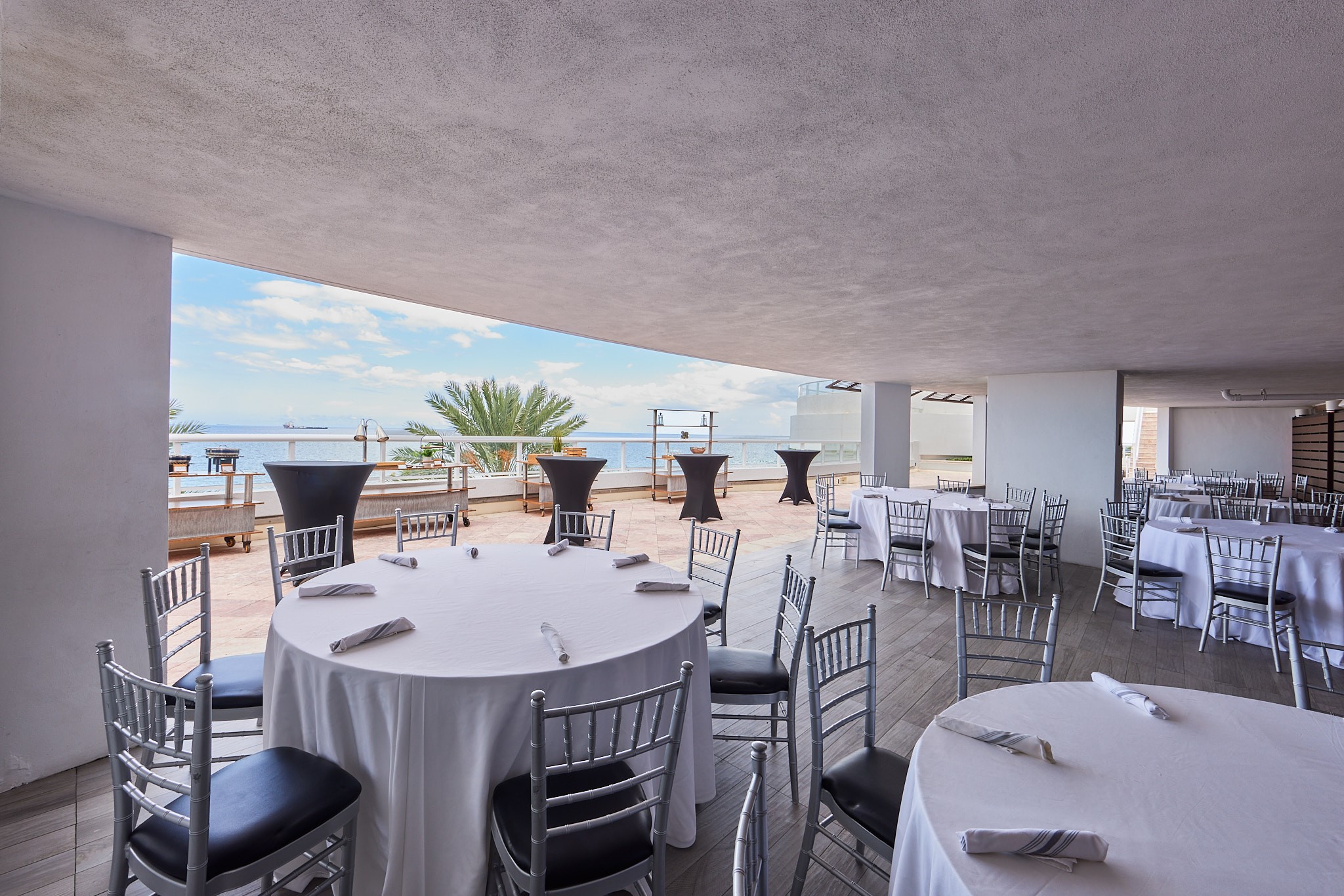 A terrace under an awning next to a beach side balcony patio, with round banquet tables topped with white linens