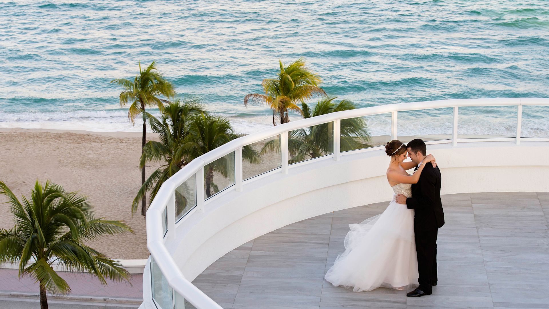 A newly married couple embrace on a patio overlooking palm trees and the ocean