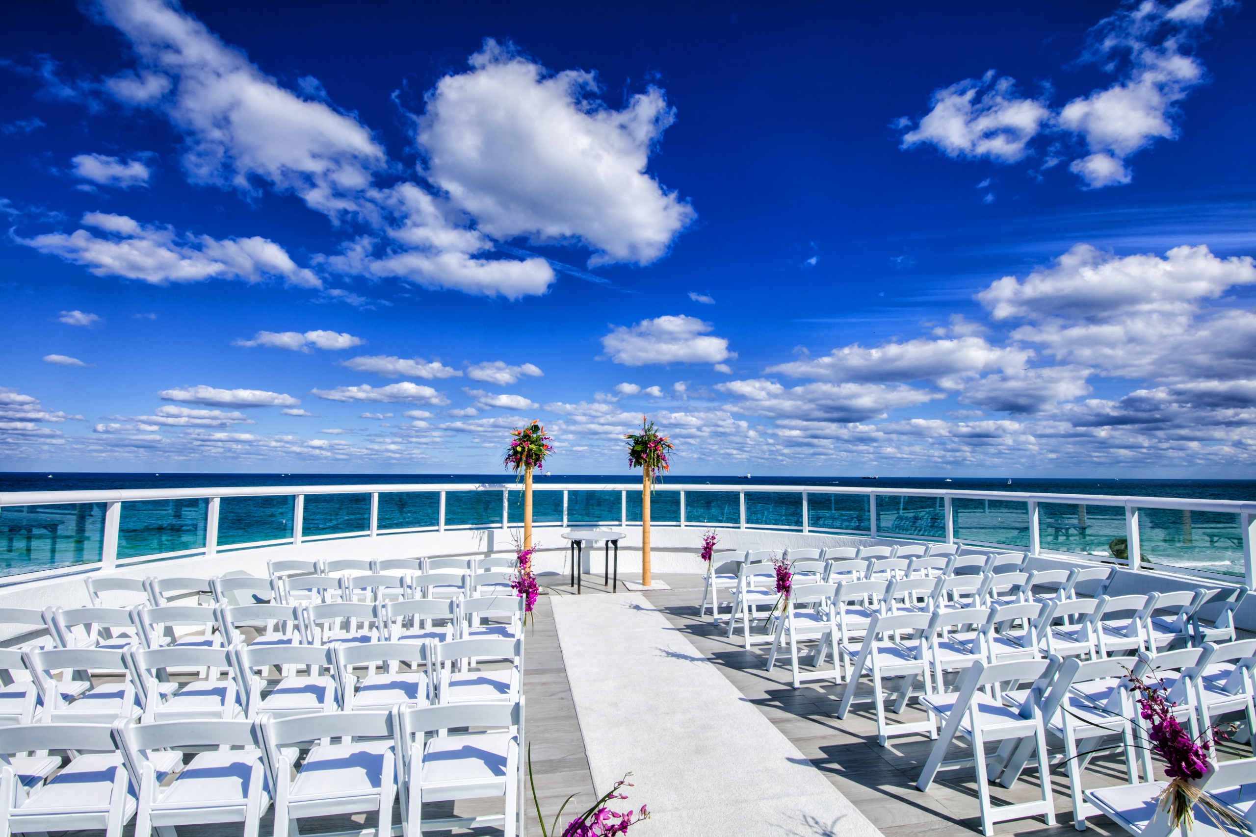 white chairs line both sides of the aisle withthe background of a brilliant blue sky over a yet to be filled wedding ceremony venue
