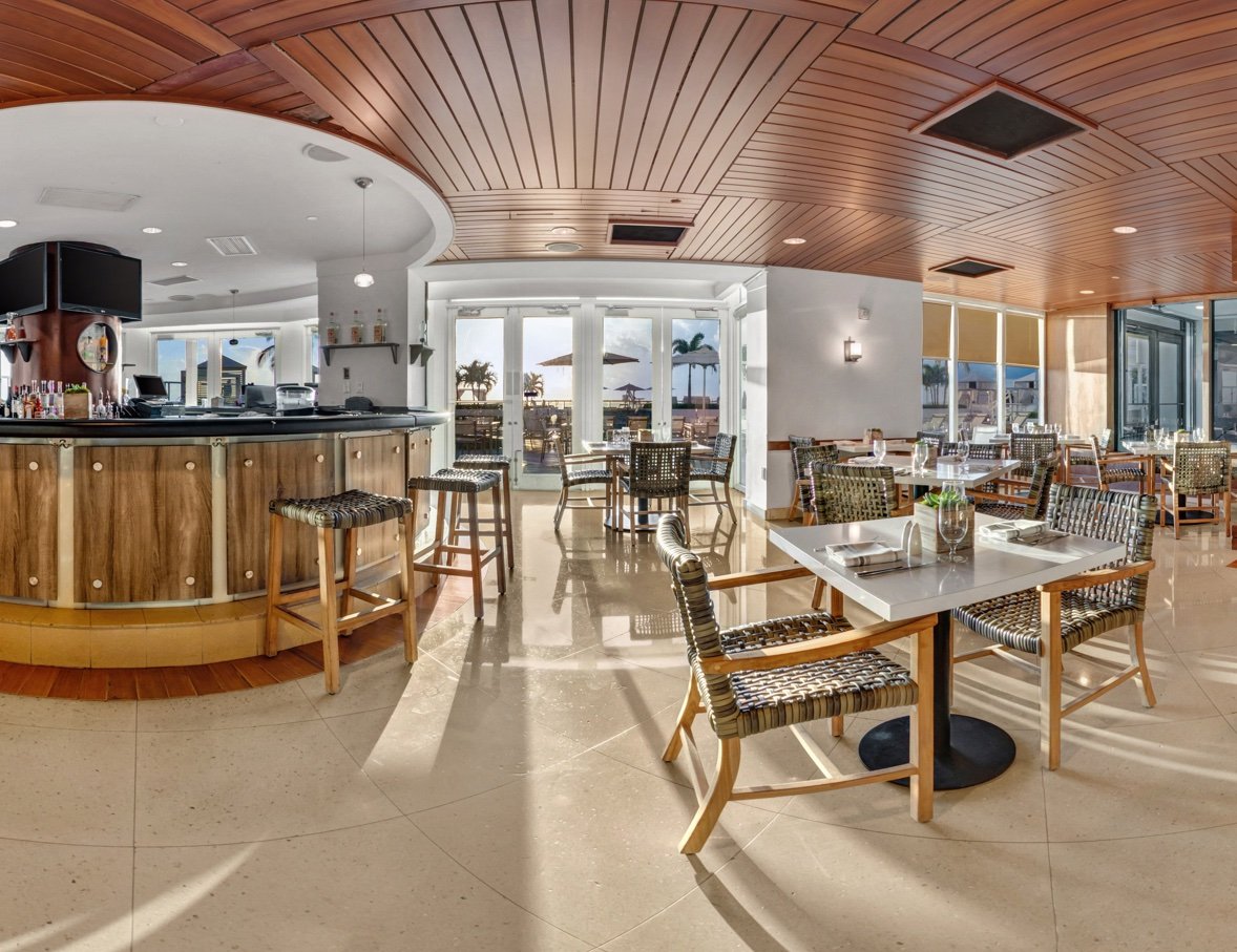Wide view of an airy restaurant in view of the beach, with a circular bar and a large dining room