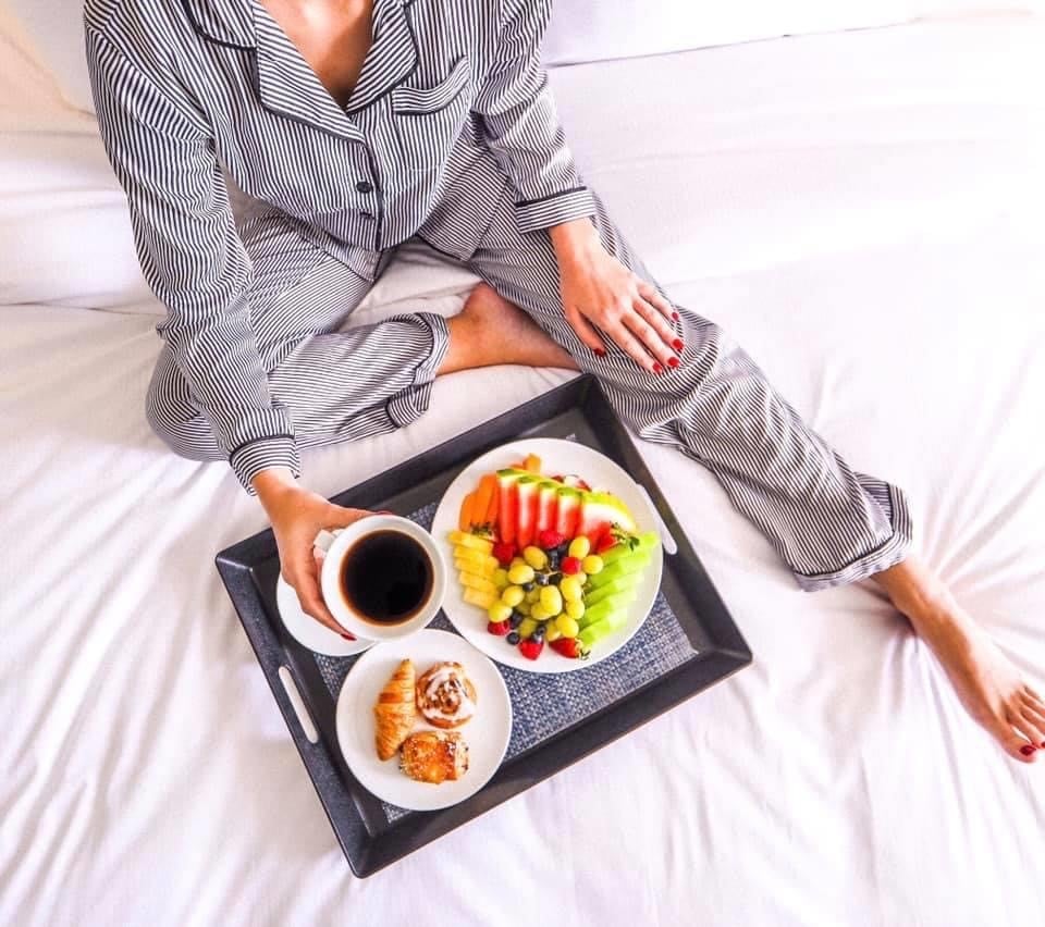 a woman in stripped pyjamas sits on a bed of white sheets with a a coffee, fruit plate and pastries